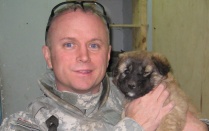 Portrait of William Mandrick, PhD, on a deployment, with a puppy found after a firefight with al Qaeda insurgents. Puppy was adopted by US forces. Mandrick observes, "We liked having dogs around because they sense when something is not right." . 