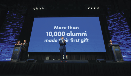 UB executives are standing in front of a blue screen with the text: "More than 10,000 alumni made their first gift.". 