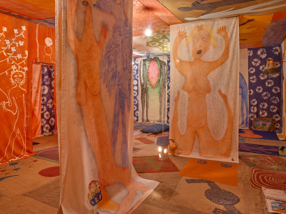 The inside of a room whose walls, floor, and ceiling are covered in brightly colored textiles of various kinds: a temple to life energies. Large floor-to-ceiling chalk-on-canvas drawings are hung around the space, imitating walls, and creating the sense of a labyrinth. Colorful cushions and small sculptures are everywhere on the floor, inviting visitors to sit or lie down to experience the work. The ceiling and floor also feature drawings of humans and animals engaged in rituals and life-giving celebrations. This is a partial view of Celina Eceiza’s installation entitled La vida terrenal reconquista al soñador which translates as Earthly Life Reconquers the Dreamer in English. 