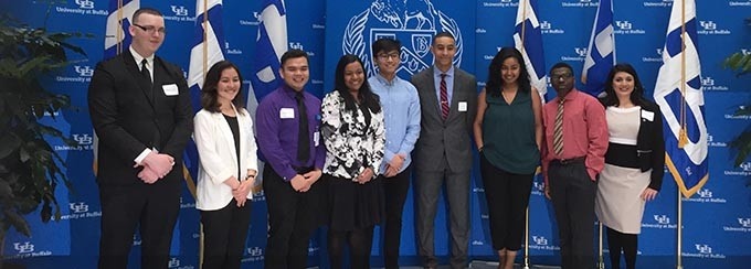 Nine students lined up in front of a blue University at Buffalo backdrop, at an awards ceremony. 