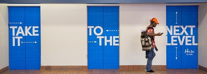 Three elevator doors closed, with the phrase "Take it to the next level" painted across each door; two words per elevator. There is a student standing to the right, looking at his cell phone. 