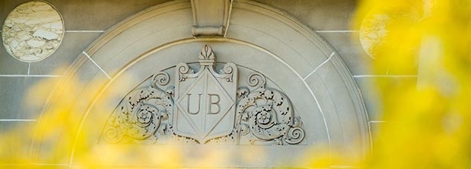 Photo of UB emblem on Foster Hall, with blurred yellow leaves framing the photo, in the foreground. 
