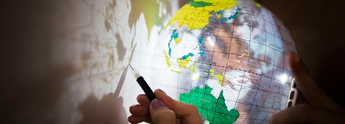 Up-close photo of a clear globe casting a shadow on a wall, and a person tracing the outline of a country with a pencil. 