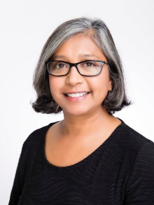 Photo of a brown woman with short grey hair, wearing rectangular black glasses and a black top - smiling at the camera. 