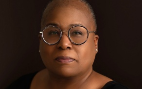 Photo shows a person in a black shirt against a black background, wearing glasses, looking into the camera. 