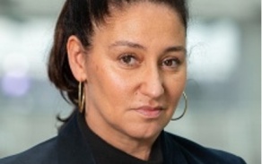 woman with dark hair tied back, hoop earrings, in a dark shirt, looking at the camera. 