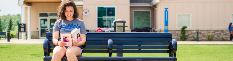 Photo is of a woman sitting outside reading a book on a bench. 