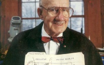man in glasses wearing a black coat over a red bow tie and white button-up shirt. 