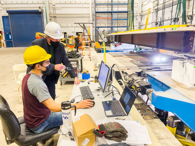 A faculty member stands and watches over a graduate student who is using two laptops to conduct modeling on a bridge. A steel beam sits in front of each of them. 