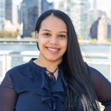 Julissa Adames-Torres smiling in a navy blue shirt on a waterfront background in front of a cityscape. 