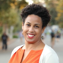 Naniette H. Coleman smiling with an orange shirt and white sweater in outdoor fraded background with trees and a walkway. 