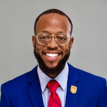 Terrance Lewis smiling in a blue blazer and red tie on a gray background. 