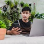 A person sitting at their laptop, looking at their phone, surrounded by plants. 