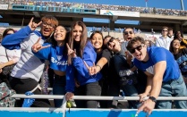 7 students wearing their best UB gear throw their "horns up" and cheer on their peers at a football game. 