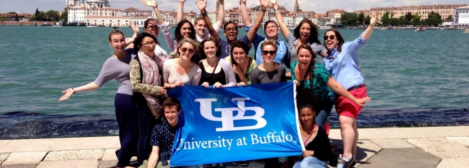 16 students post with a royal blue "UB" flag in front of a canal in Venice, Italy on a trip with the Student Leadership International Dialogue and Exchange (SLIDE) Program. 