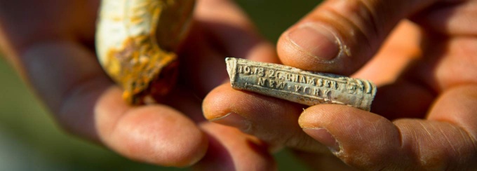 Close-up of hands holding small historical artifacts, including a stamped fragment labeled &ldquo;New York,&rdquo; representing archaeological research. 