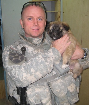 Portrait of William Mandrick, PhD, on a deployment, with a puppy found after a firefight with al Qaeda insurgents. Puppy was adopted by US forces. Mandrick observes, "The combat out-posts liked liked having dogs around because they sense when something is not right.". 