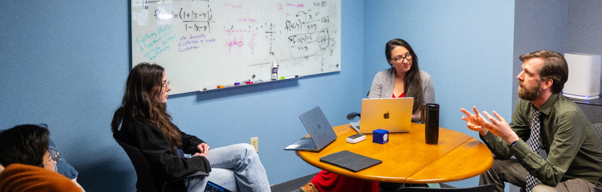Four people sit around a small round table in a blue-walled office, engaged in discussion. A man gestures as he speaks while two women with laptops listen, and a whiteboard behind them is filled with equations, diagrams, and notes. 