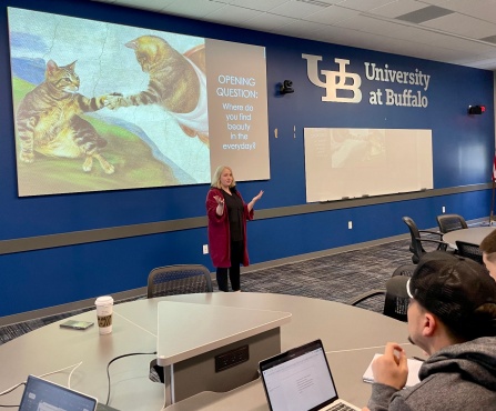 Sarah Vincent, associate teaching professor of philosophy, stands in a classroom with a slide behind her that asks, "Where do you find beauty in the everyday?". 