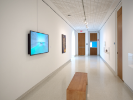 An installation image at UB Anderson Gallery of a hallway. In the foreground is a bench, and visible on the wall is a screen showing a blue vortex. 