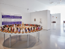 An installation photograph of Thinking in Indian at UB CFA Gallery. In the foreground is a table where cornhusk dolls are arranged in a ceremonial-looking circle. This is a work by Elizabeth Doxdater. 