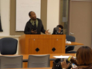 Man presents at a podium while audience in the classroom watches