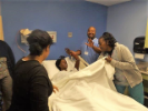 Woman holds blanket up while man holds training mannequin's arm and others look on around the hospital bed