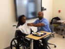 Man prepares to feed woman in a wheelchair from a hospital tray in a classroom