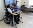 Man demonstrates how to feed woman in a wheelchair from a hospital tray in a classroom
