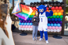 A student with UB Mascot Victor E. Bull taking a picture in front of a display of rainbow balloons, and holding a progress pride flag. Photographer: Meredith Forrest Kulwicki