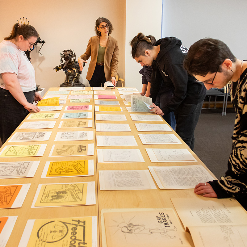 A group of students and instrctor stand around a large table covered with neatly arranged printed pages in various colors. One person near the center gestures toward the materials while others lean over and closely examine individual sheets.