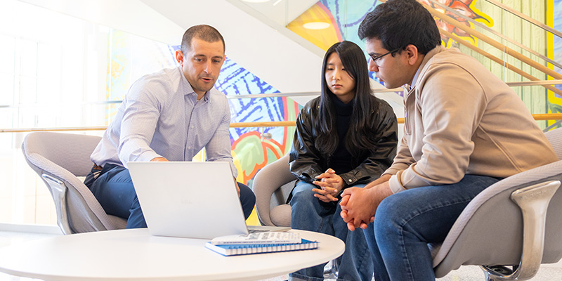 Three people sit around a small round table in a bright, modern indoor space, looking at an open laptop. One person gestures toward the screen while the other two lean forward with hands clasped, focusing on the laptop.