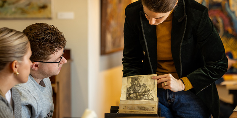 A person holds open an old illustrated book, pointing to a detailed black-and-white image on one page, while two others sit nearby and look closely. The scene takes place in a softly lit indoor setting with artwork on the walls, and the book rests above a small stand or stack beneath it.