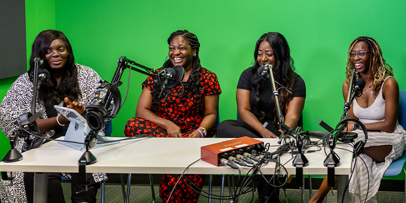 Four people sit at a table in front of a green screen backdrop, speaking into microphones on adjustable arms. Audio equipment and cables are spread across the table, including a small red interface unit, while one person gestures toward a tablet and others face their microphones during the recording setup.