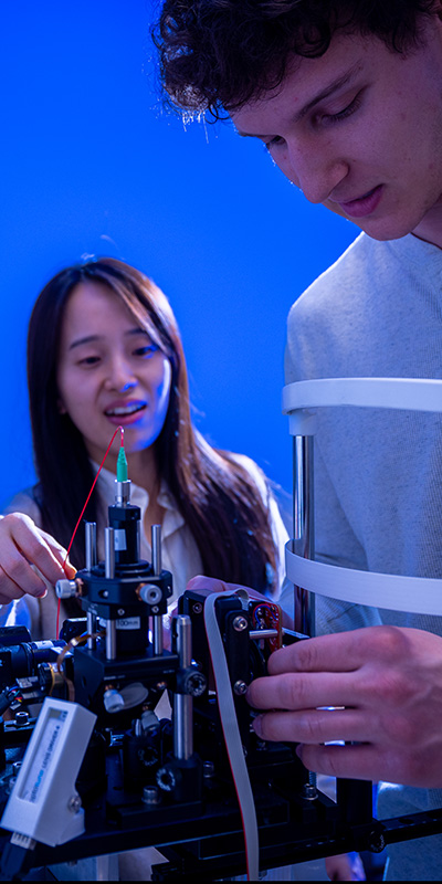 Two people work together on a compact laboratory device under blue lighting. One person adjusts a small component on the machine with both hands, while another points toward a needle-like part connected by a thin red wire. The setup includes metal supports, a syringe-like element, and electronic components arranged on a small platform.