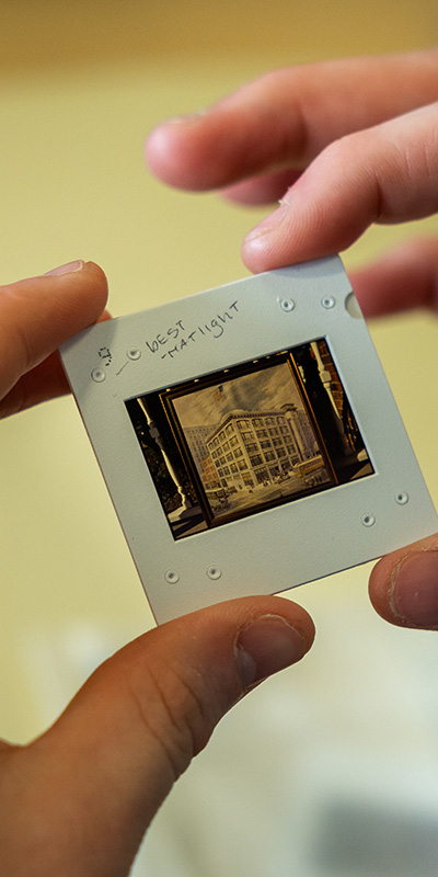 A close-up view shows two hands holding a photographic slide in a white mount. The small transparent frame reveals an image of a multi-story building with windows and architectural details. Handwritten text is visible on the slide mount, and the background is softly blurred in warm tones.