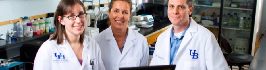 Three people stand in front of a laptop, in a lab, wearing white coats. 