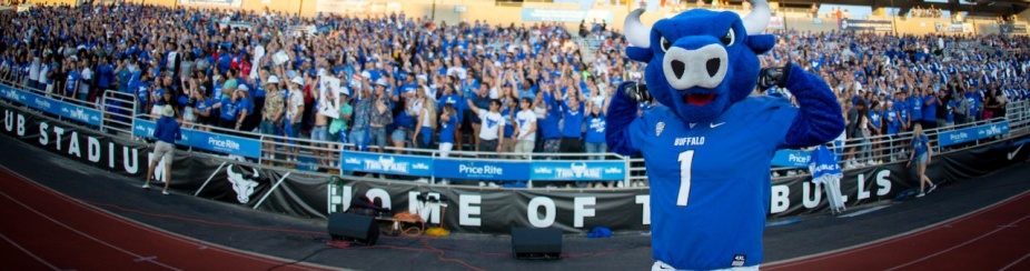UB's mascot Victor E. Bull makes two large bicep muscles while standing in front of a packed stadium at a UB sporting event. 