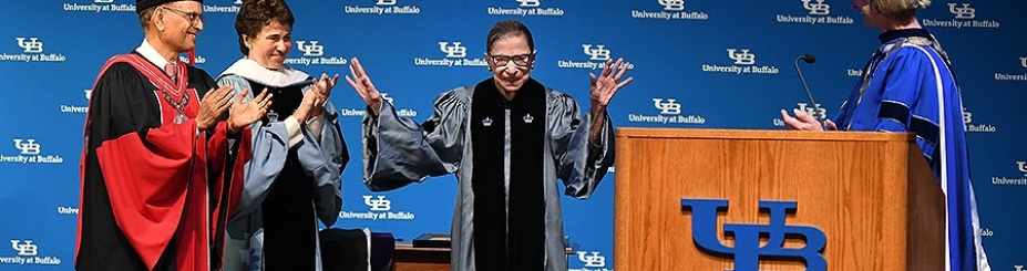 Justice Ruth Bader Ginsberg motions to the audience to sit down as she stands on stage near the podium with UB president Tripathi and other UB staff. 