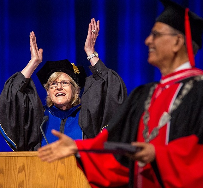 Robert Schulze wears a cap and gown at a podium with her hands in the air. President Tripathi is blurred in the foreground. 