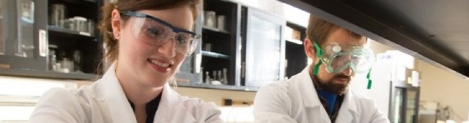 Two students wearing rubber gloves and goggles, working in a lab environment. 