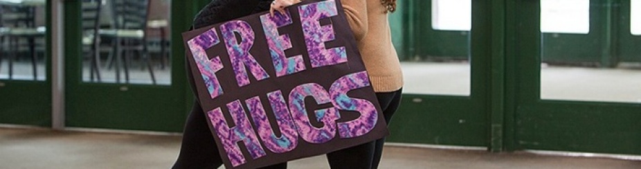 Two students embrace in the student union lobby while one holds a sign that reads "free hugs". 