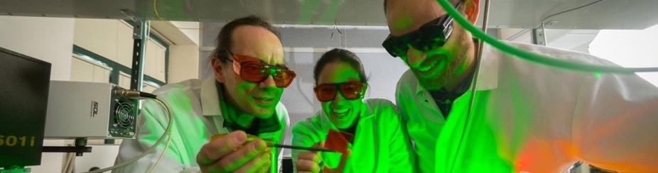 Three people look at a specimen in a chemistry lab with a green light shining on their faces. 