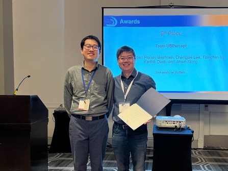 Jinjun Xiong is holding the UB Percept team's TinyML competition certifcate and standing next to competition organizer Zhenge Jia at the ICCAD conference. Behind them is a projection screen displaying the names of the UB Percept team. 