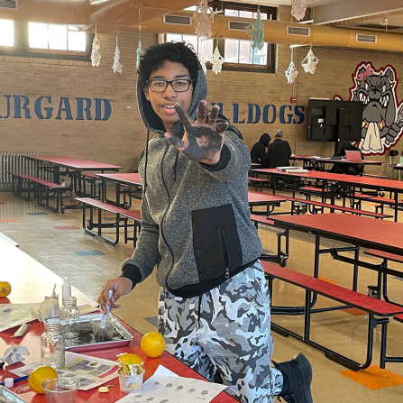 A student wearing a hoodie and pants kneels with one knee on a cafeteria bench. His hand is smudged with ink and a finger printing kit sits on the table in front of him. He is learning forensics.