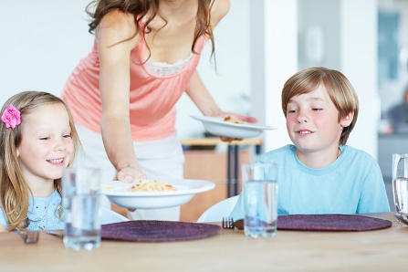 A mother places big bowls of macaroni and cheese before a boy and a girl sitting at the table.