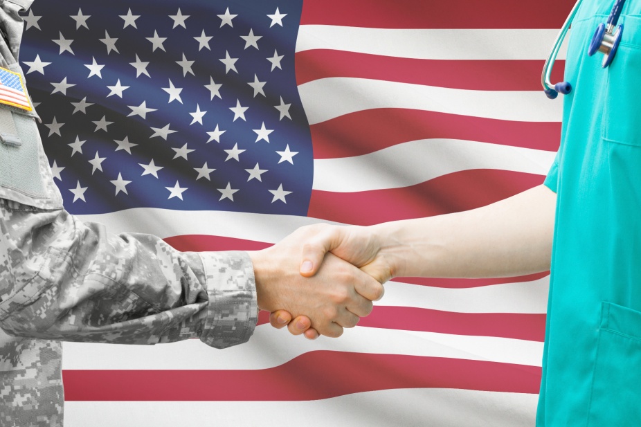 A service member and a health care practitioner shake hands in front of the American flag. 