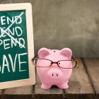 Photoillustration of the concept of saving featuring a piggybank wearing glasses and a chalkboard with the words "spend, spend, spend" crossed out and "save" written below. 