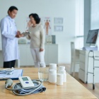 Doctor and patient in background; in foreground is rtable containing pill bottles, glass of water, chart and stethoscope and blood pressure cuff. 