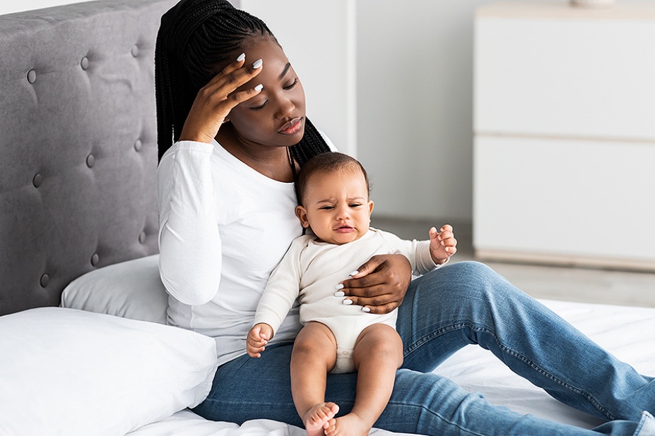 Black mother sitting on bed with child, looking overwhelmed. 